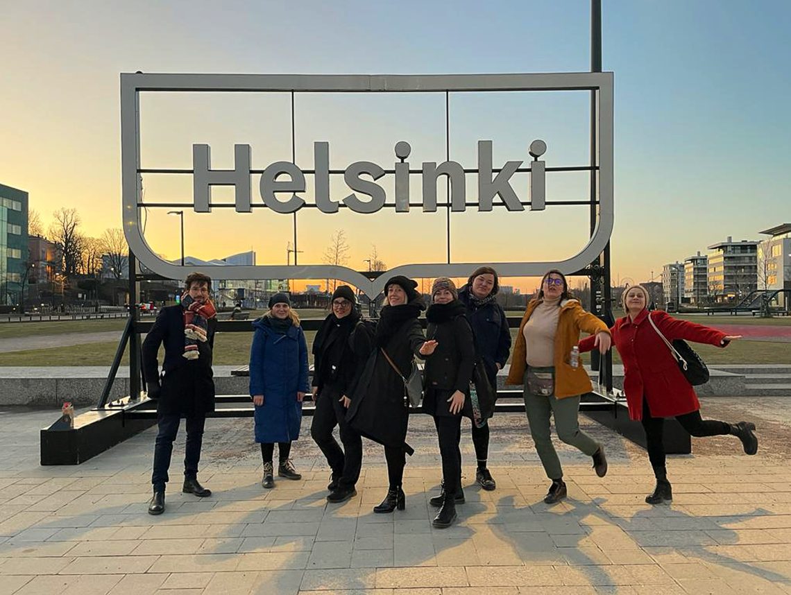 Zdjęcie przedstawia grupę osób z wyjazdu na tle napisu "Helsinki" i zachodzącego słońca.