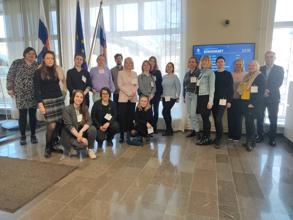 grupa wyjazdowa wraz przedstawicielami fińskich ministerstw i NGOsów pozuje na tle flag Finlandii, UE i Ukrainy.