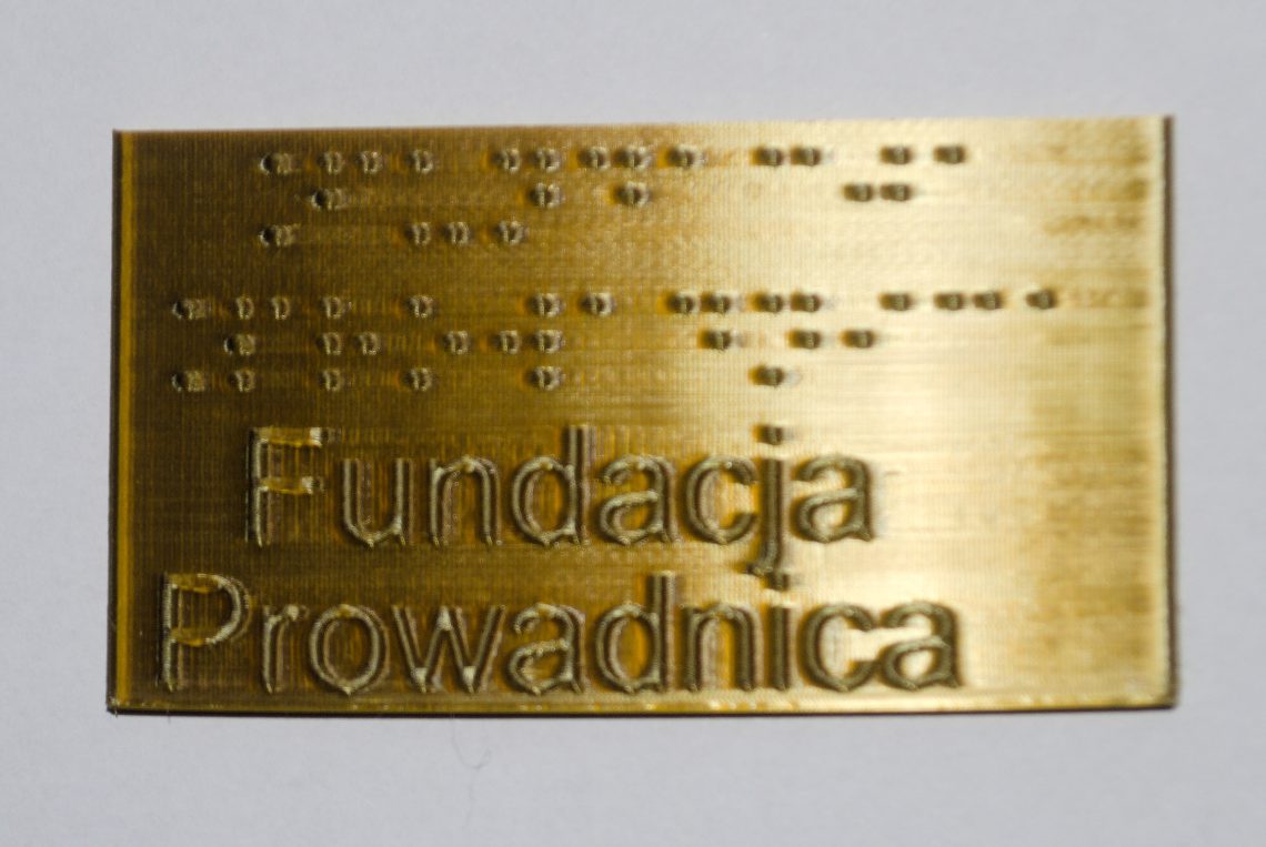 Mosiężna tabliczka z nazwą Fundacji Prowadnica zapisaną alfabetem Braille'a oraz alfabetem łacińskim