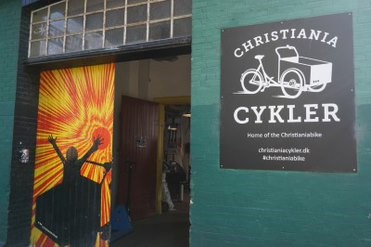 wejście do warsztatu rowerowego w zielonym budynku. Na wejściu żółto-pomarańczowo-czarna grafika z dzieckiem w przyczepce rowerowej. Na ścianie czarna tablica z grafiką roweru i napisem "Christiania cykler"