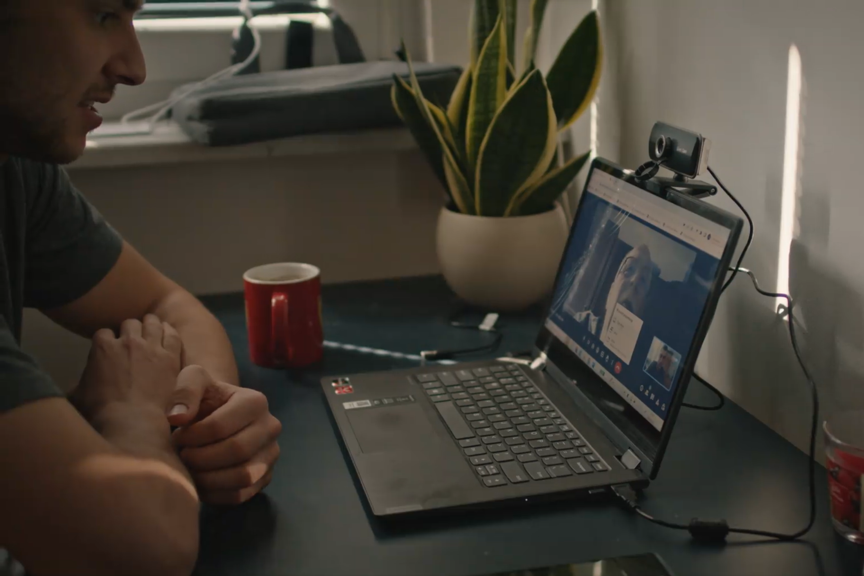 Kadr z filmu E-care. Na zdjęciu mężczyzna (asystent) mówi do kamery internetowej podłączonej do laptopa. Na ekranie laptopa widać jego klienta, starszego mężczyznę.