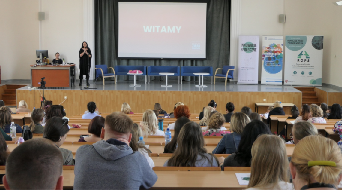 sala wykładowa - widok z sali na podwyższenie dla prowadzących, uczestnicy widoczni od tyłu, na ekranie napis "witamy"