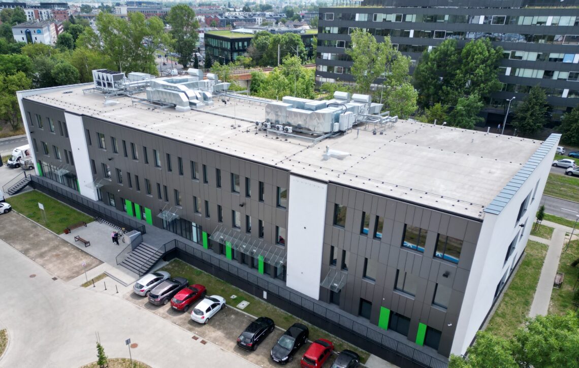 widok z lotu ptaka na budynek biurowy, szara fasada z białymi pionowymi pasami oraz jasnozielonymi elementami na parterze, przed budynkiem fragment parkingu, w tle widok na okolicę, drzewa i budynki mieszkalne, nowe i stare