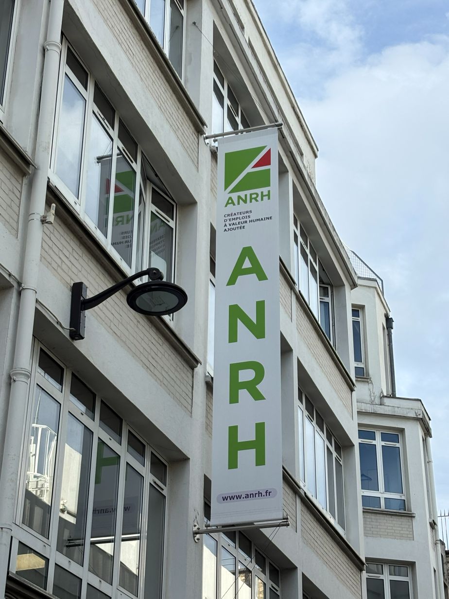 pionowy szyld z napisem w kolorze zielonym ANRH, przymocowany do ściany budynku o dużych oknach