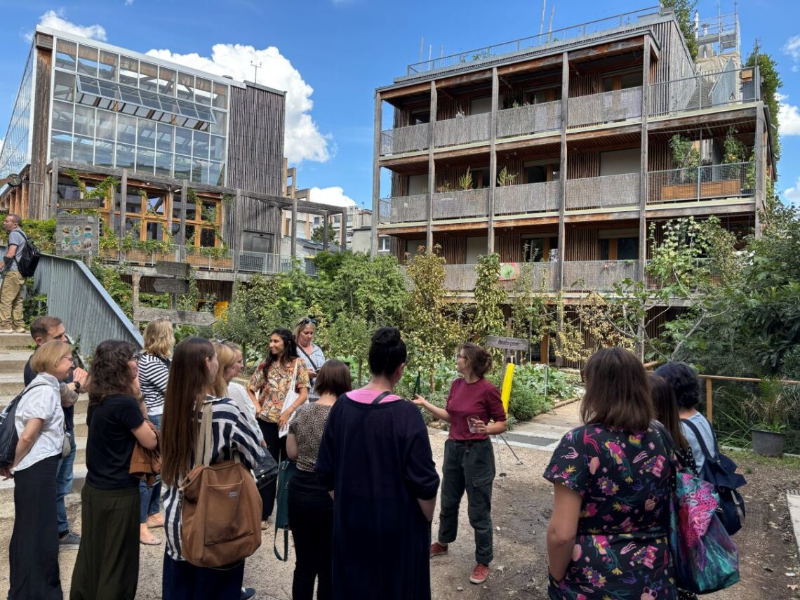 grupa osób z przewodnikiem na tle dwóch betonowych budynków, na dalszym planie widoczne krzewy i inne rośliny
