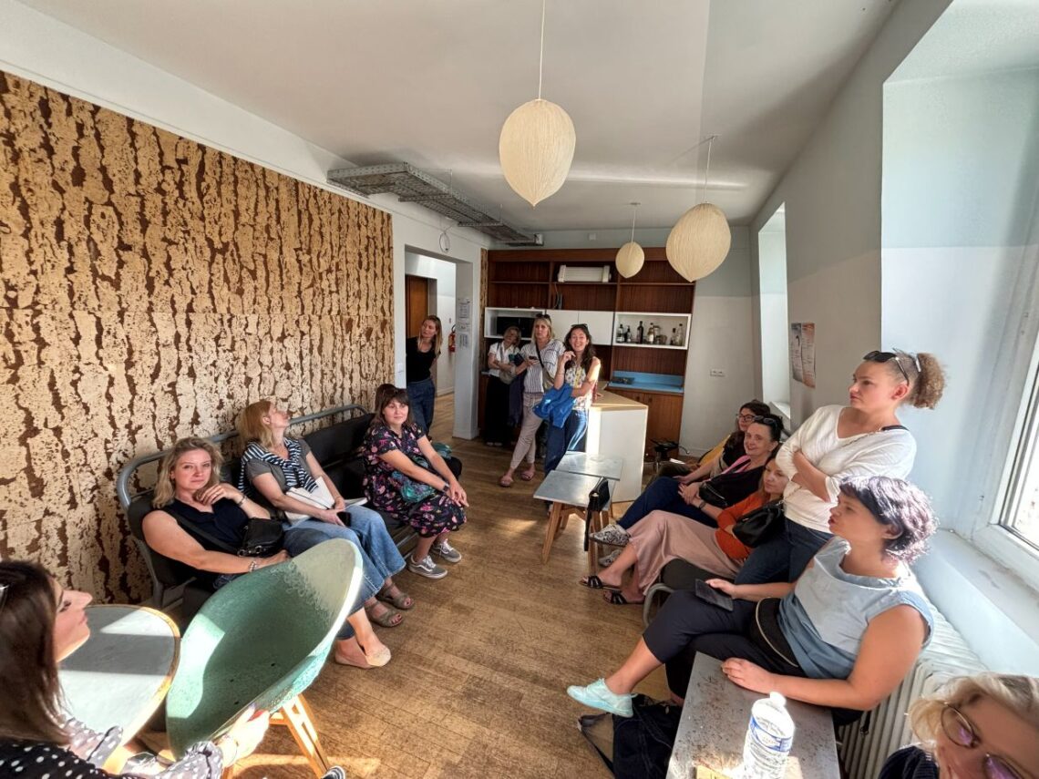 grupa osób siedząca po obu stronach korytarza, na lewej ścianie duży panel w nieregularne brązowo-czarne pasma