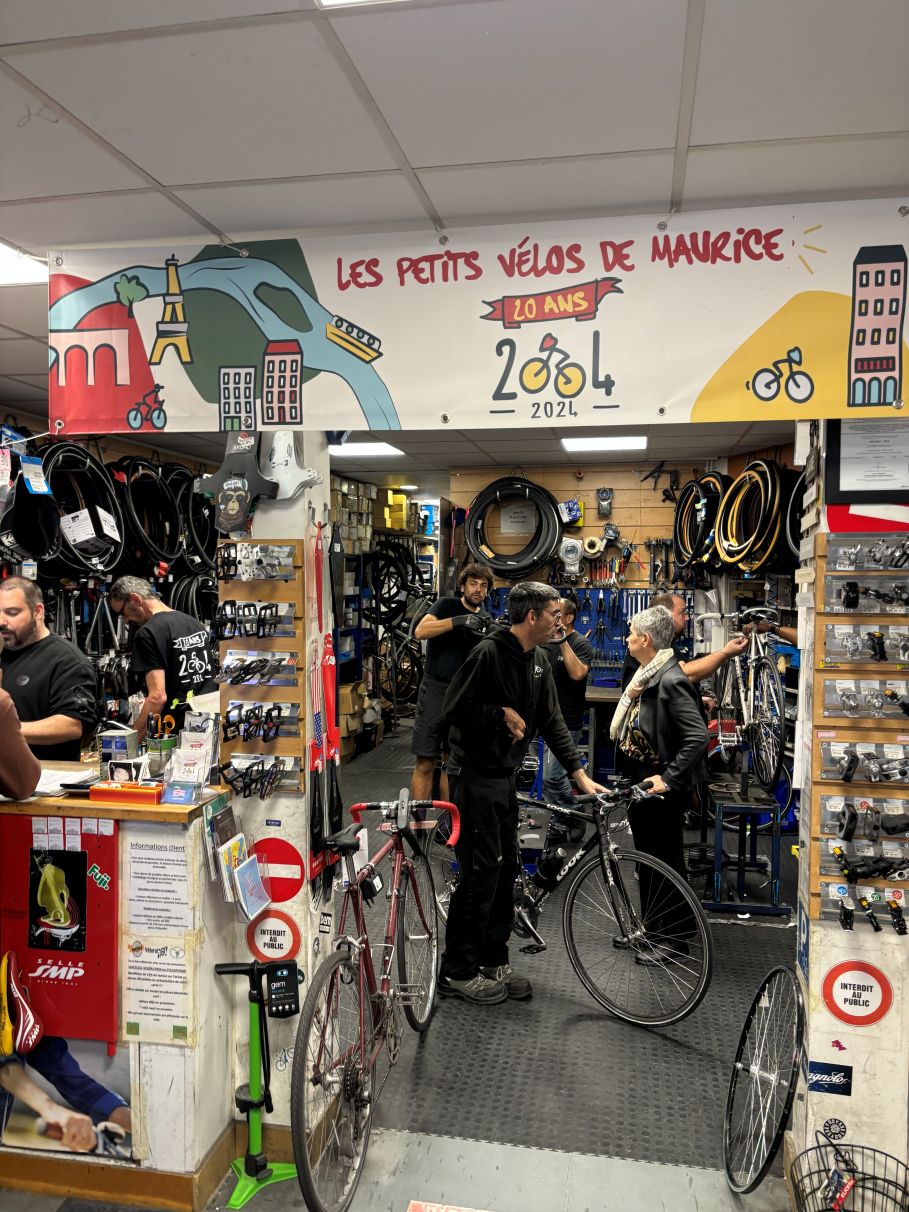 wąskie wejście do warsztatu rowerowego, przy wejściu półki z różnymi akcesoriami, nad wejściem napis po francusku "les petits velos de Maurice"