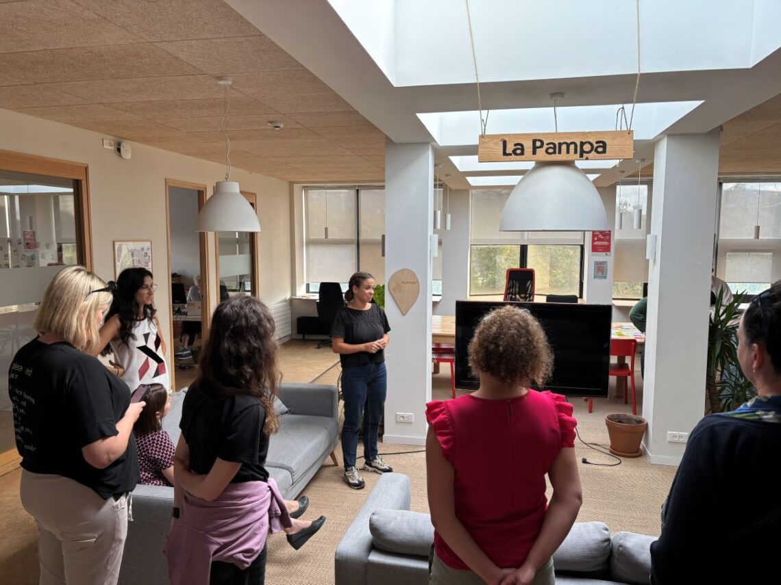 kobieta opowiadająca coś grupie osób w pomieszczeniu, jasnobeżowe kolory ścian, białe kolumny i abażur