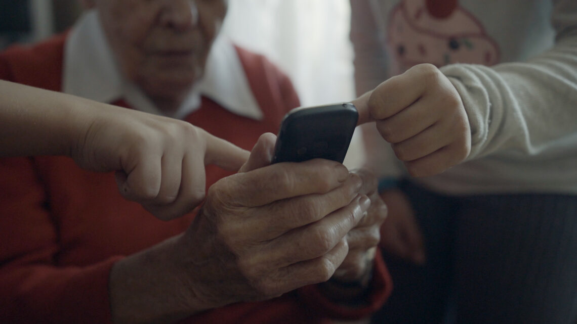 Seniorka trzyma telefon, dziewczynki wskazują jej coś palcem na ekranie.