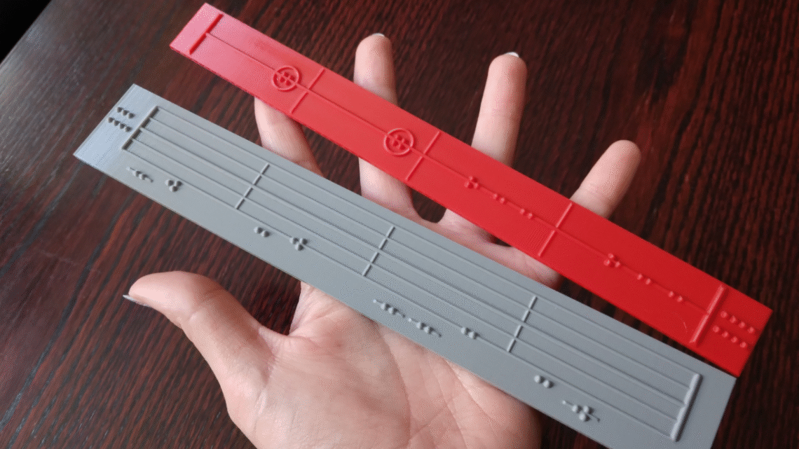 dłoń, na niej dwa podłużne elementy z wytłoczonym zapisem nutowym dla osób niewidomych