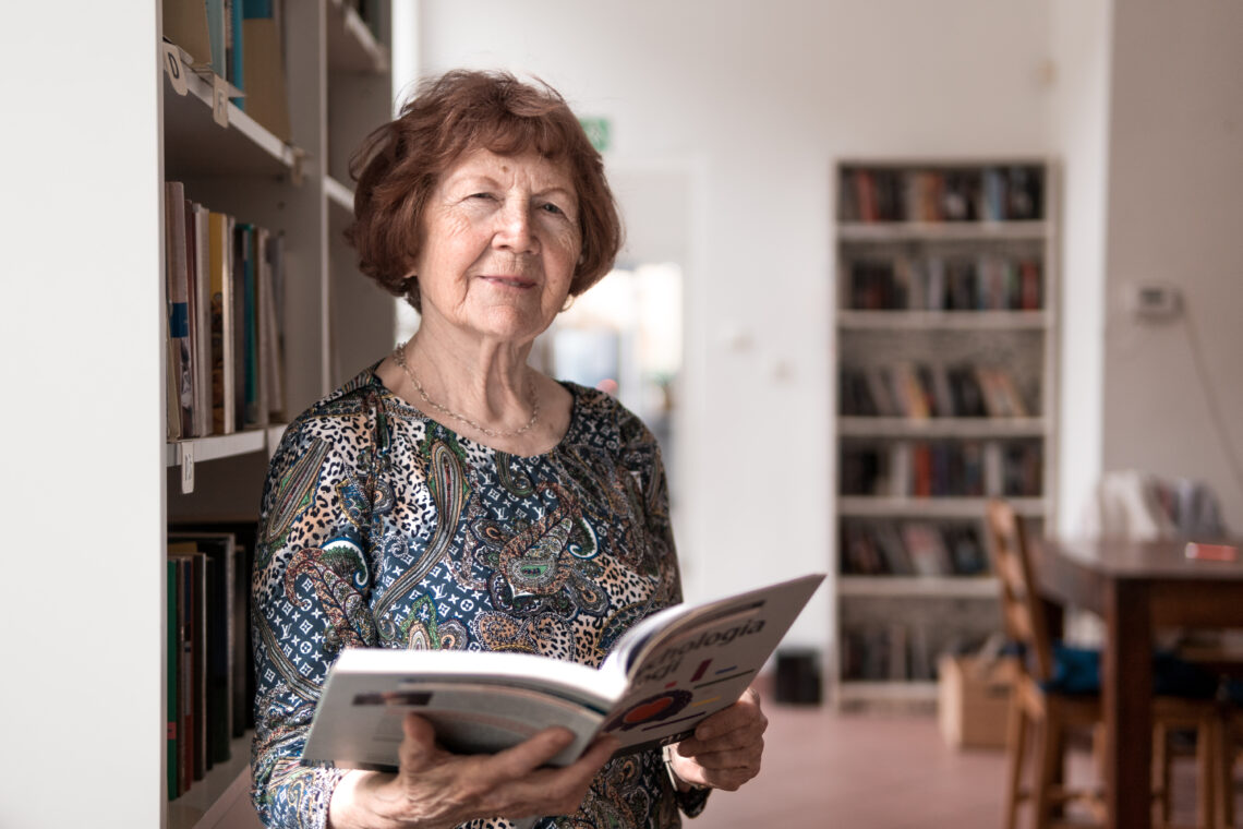 starsza kobieta z książką w rękach na tle szafy bibliotecznej