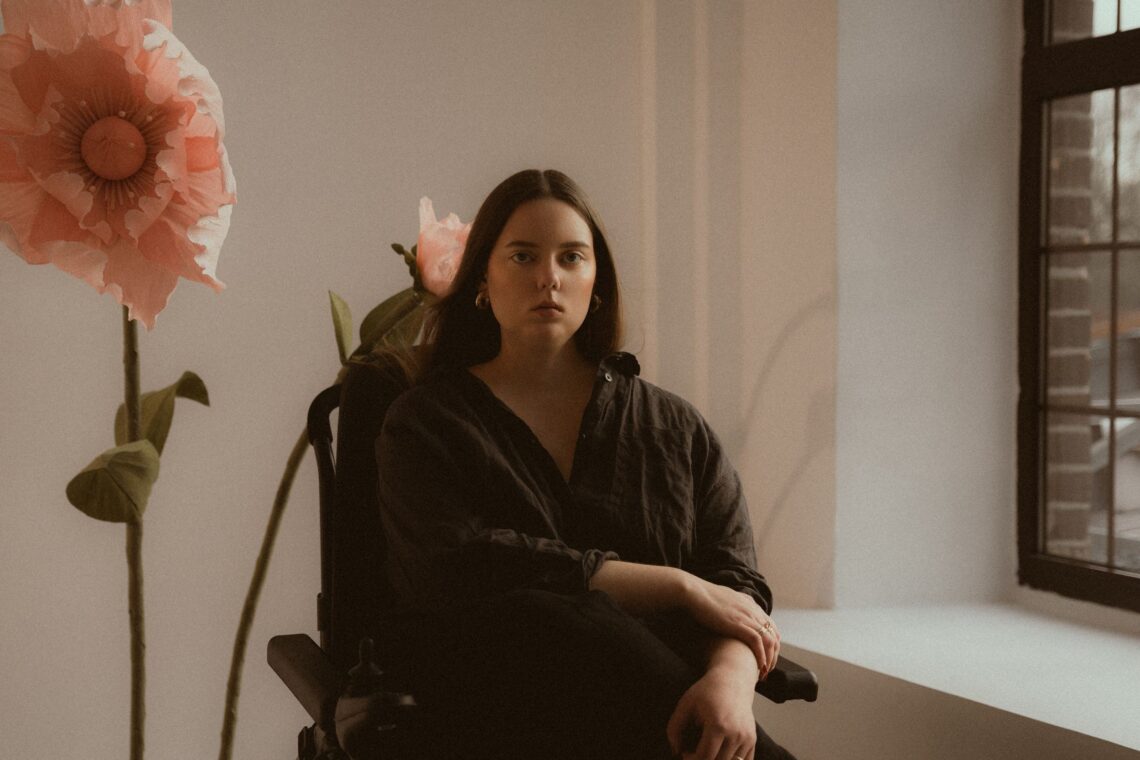 młoda kobieta siedząca na wózku, po jej lewej stronie okno, po prawej duży różowy kwiat