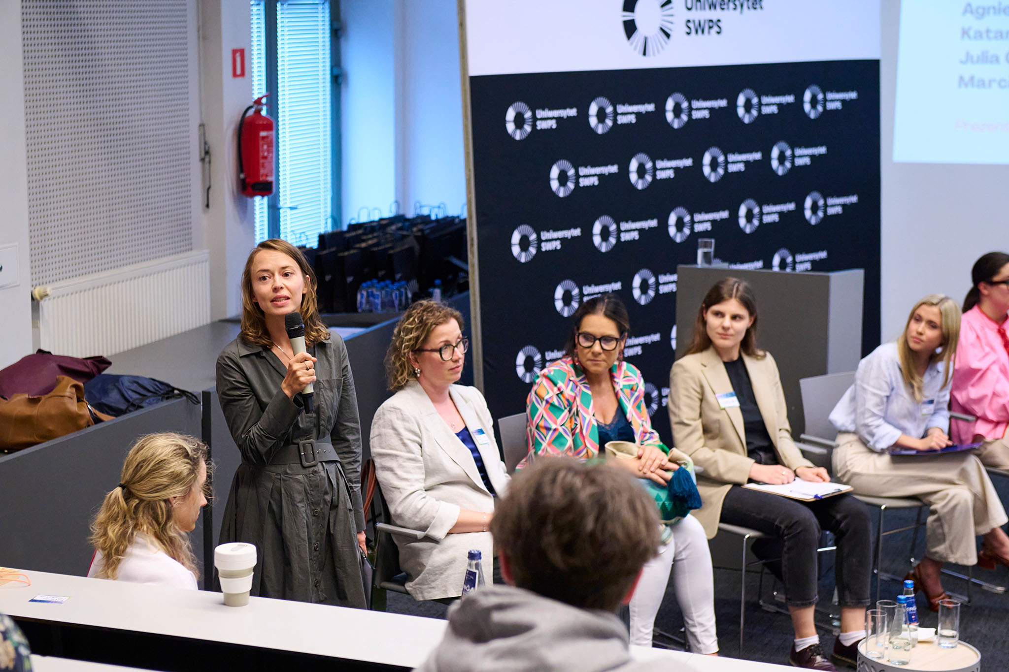 Kobieta trzyma mikrofon i zabiera głos podczas panelu dyskusyjnego. Obok niej siedzi kilka uczestniczek wydarzenia, a na pierwszym planie widoczni są słuchacze. Spotkanie odbywa się w sali konferencyjnej z widocznym w tle logo Uniwersytetu SWPS oraz ekranem prezentacyjnym.