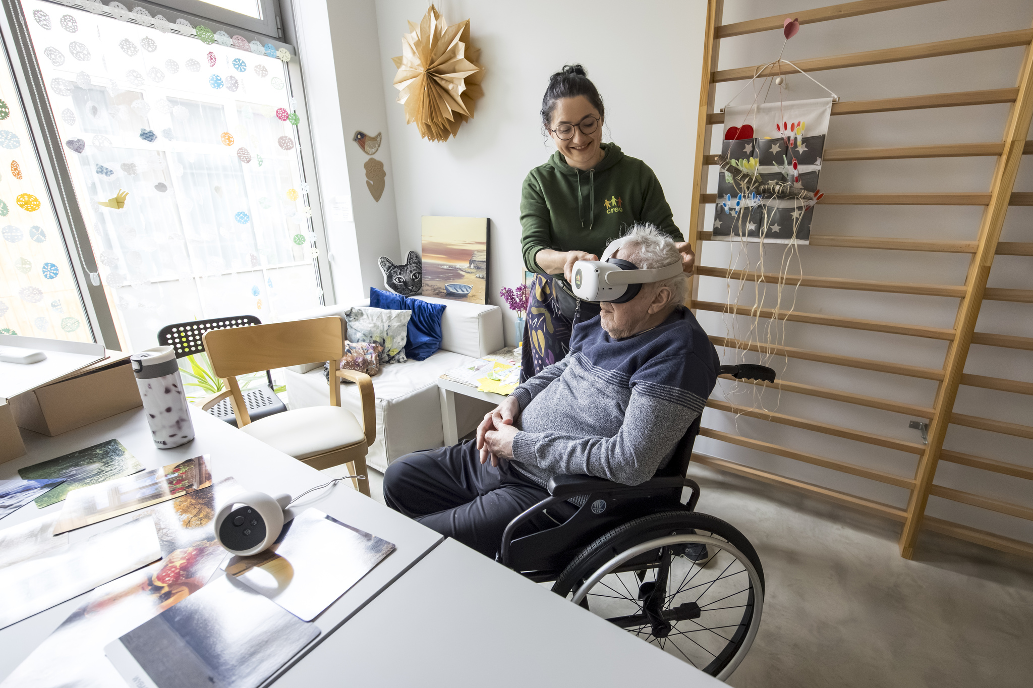 Animatorka wraz z seniorem w trakcie zajęć z użyciem gogli vr. Senior siedzi na wózku inwalidzkim, ma założone gogle vr, za nim stoi animatorka , uśmiecha się i pomaga dopasować gogle