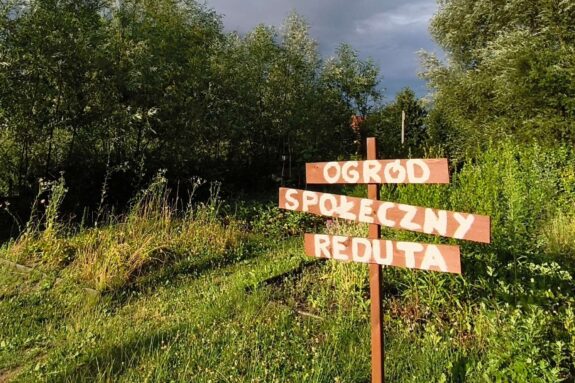 Drewniana tabliczka z napisem "Ogród społeczny Reduta" na tle terenu zielonego i lasu