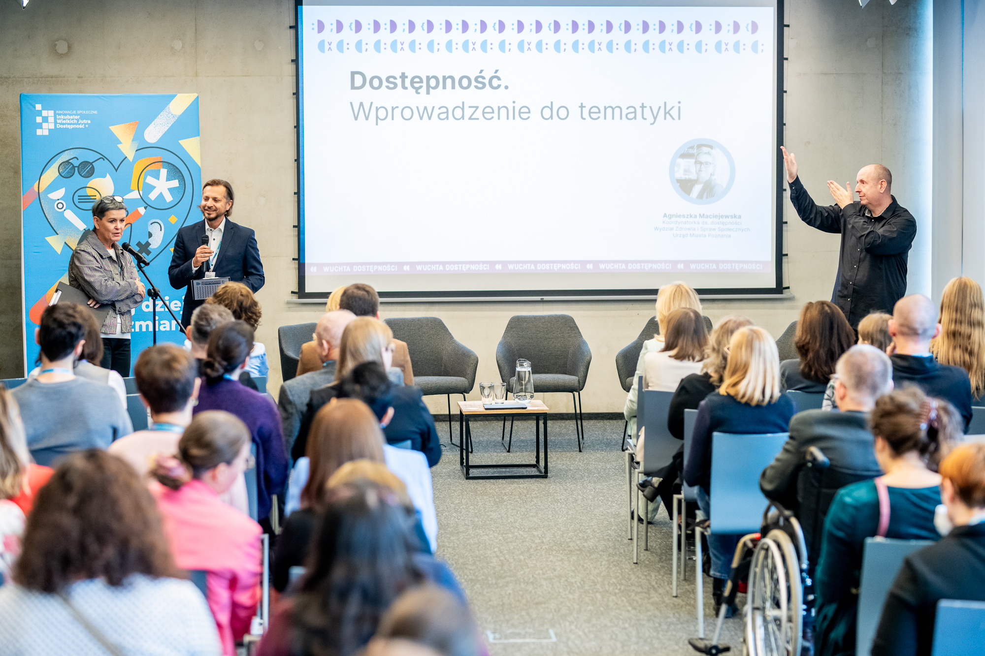 Konferencja dotycząca dostępności zdjęcie obejmuje salę, ludzie siedzący na krzesłach widoczni są plecami, na ekranie wyświetlana jest prezentacja