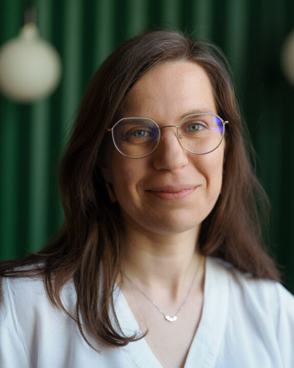 Portret uśmiechniętej kobiety z długimi brązowymi włosami i okrągłymi okularami, ubranej w białą bluzkę.