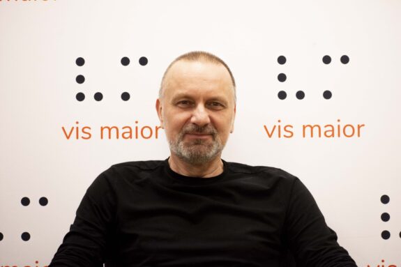 Uśmiechnięty Pan z siwym zarostem na tle Loga Fundacji Vis Maior, gdzie nad nazwą jest grafika V i M z alfabetu braille'a