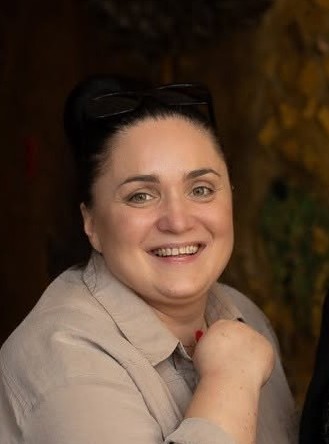Uśmiechnięta kobieta z ciemnymi włosami, ubrana w jasną koszulę. Na głowie ma okulary