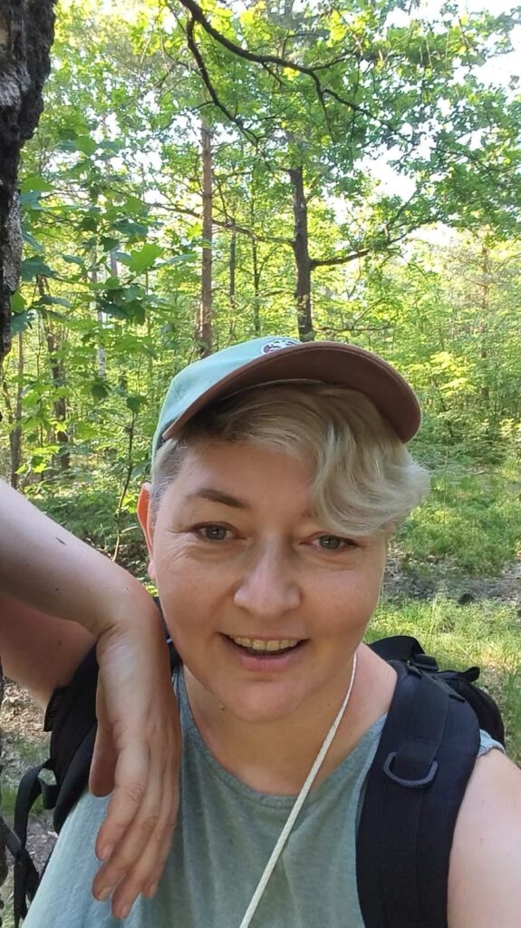 Blond kobieta w czapce z daszkiem, uśmiecha się oparta o drzewo w lesie.
