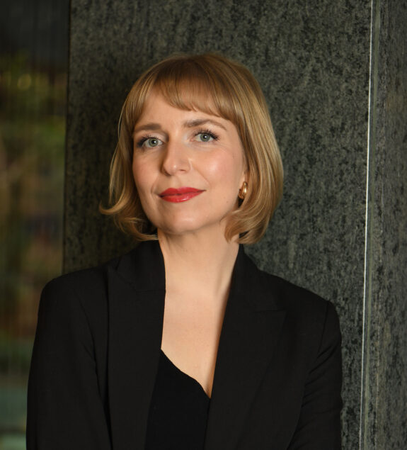 Portret kobiety z krótkimi, jasnobrązowymi włosami i grzywką, ubranej w czarną marynarkę.