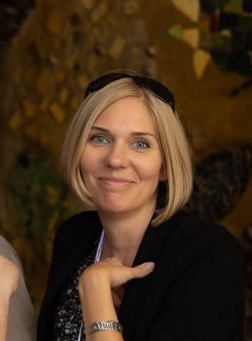 Uśmiechnięta kobieta z blond włosami, ubrana w czarną marynarkę. Na głowie ma okulary