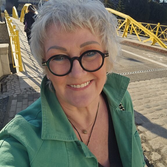 Uśmiechnięta kobieta w dużych, okrągłych okularach i zielonej kurtce robi selfie na tle żółtego mostu