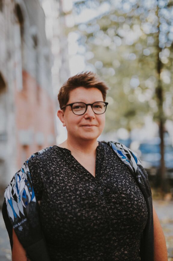 Portret uśmiechniętej kobiety na ulicy z krótkimi, brązowymi włosami w czarnej koszuli i okularach