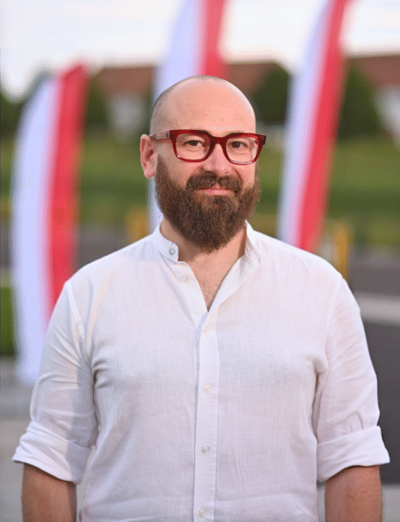 Łysy mężczyzna z bujnym zarostem w okularach o czerwonych oprawkach, ubrany w białą koszulę na tle polskich Flag pionowych.